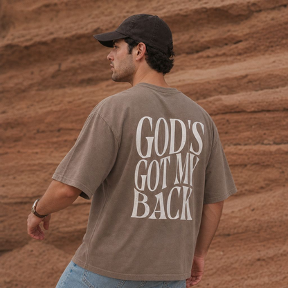 Man wearing taupe oversized graphic t-shirt with “God’s Got My Back” faith design on back in desert landscape setting